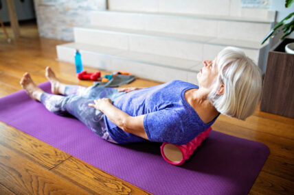 senior woman stretching her back on a foam roller, lying down at on an exercise mat at home